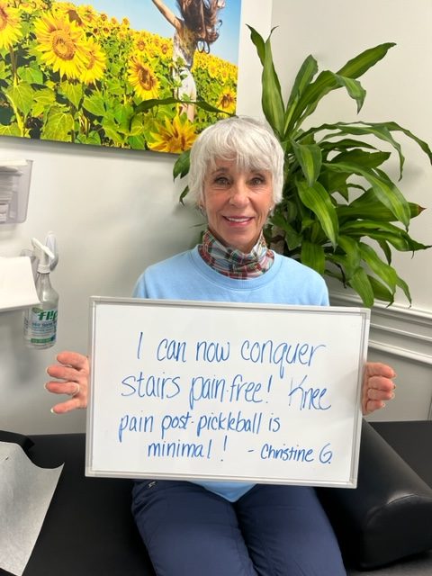 Christine G., a Vitality Healing Center patient, holding a handwritten whiteboard that reads: I can now conquer stairs pain free. Knee pain post-pickleball is minimal.