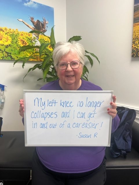 Susan R., a Vitality Healing Center patient, holding a handwritten whiteboard that reads: My left knee no longer collapses and I can get in and out of a car easier.