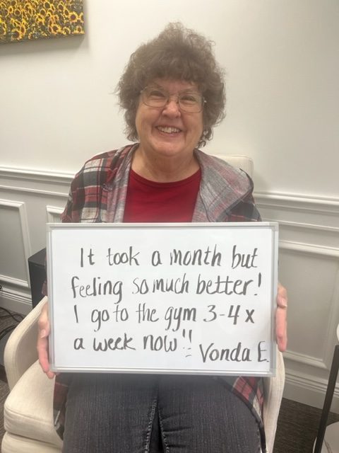 Vonda E., a Vitality Healing Center patient, holding a handwritten whiteboard that reads: It took a month but feeling so much better. I go to the gym 3 to 4 times a week now.