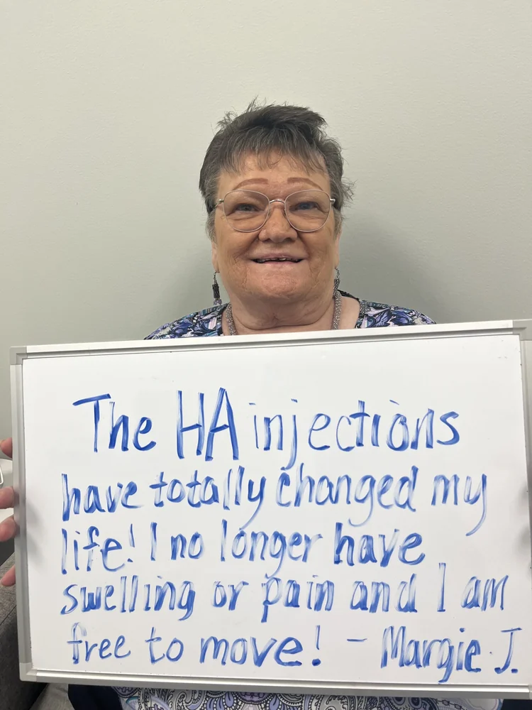 Margie J., a Vitality Healing Center patient, holding a handwritten whiteboard that reads: The HA injections have totally changed my life. I no longer have swelling or pain and I am free to move.