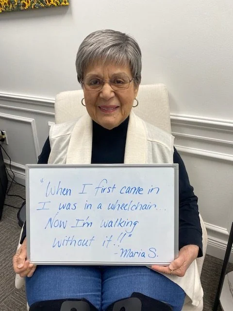 Maria S., a Vitality Healing Center patient, holding a handwritten whiteboard that reads: When I first came in I was in a wheelchair. Now I'm walking without it.