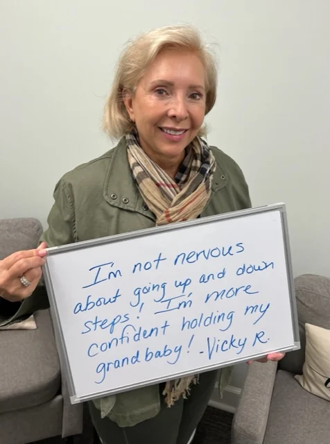 Vicky R., a Vitality Healing Center patient, holding a handwritten whiteboard that reads: I'm not nervous about going up and down steps. I'm more confident holding my grand baby.