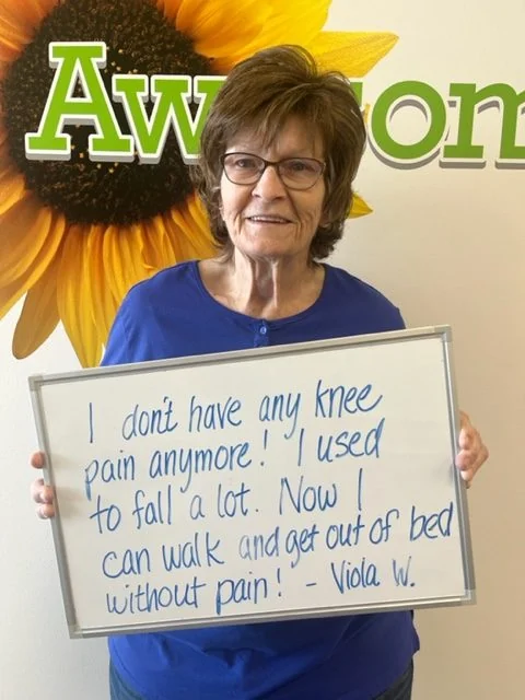 Viola W., a Vitality Healing Center patient, holding a handwritten whiteboard that reads: I don't have any knee pain anymore. I used to fall a lot. Now I can walk and get out of bed without pain.