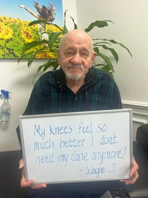 Wayne J., a Vitality Healing Center knee pain patient, holding a handwritten whiteboard that reads: My knees feel so much better I don't need my cane anymore.